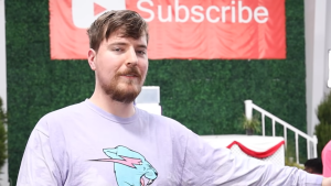 MrBeast stood wearing a light purple jumper in front of a green foliage wall with a YouTube banner behind him which says 'Subscribe.'