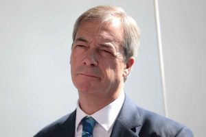 Close up image of Nigel Farage. Can see his face, staring into the distance, and the top of his black suit with blue floral tie.