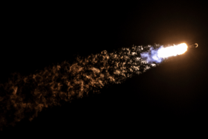 A rocket launching into space at night, leaving a bright plume of flame and exhaust smoke trailing behind it, glowing with shades of orange, blue, and white against the dark sky. SpaceX launches lunar landers Blue Ghost and Resilience on historic moon missions