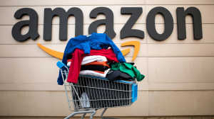 A photo of the Amazon logo with a shopping cart in front of it stacked full of clothes. The clothes include a blue shirt, a red shirt, a black shirt, and a green shirt. The shopping cart is placed on the ground. The background is a beige wall.