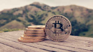 A photo of a close-up shot of a stack of Bitcoin coins. The coins have a Bitcoin symbol on them. The stack is placed on a wooden surface. In the background, there are California hills with greenery. The overall image has a soft focus.