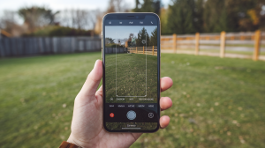 A photo of a phone screen with a camera app open. The camera is focused on a grassy field. There is a person's hand touching the phone screen. The background contains a wooden fence and some trees. The lighting is bright.