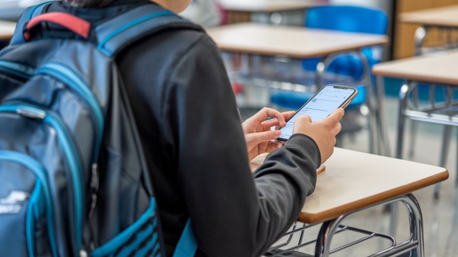 A photo of a student's back wearing a backpack, sitting in a classroom. The student is holding a phone and typing a text message. The background contains desks and chairs.