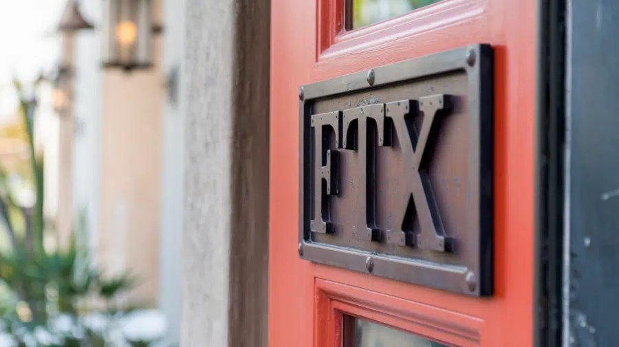 A close-up shot of a building door with the text "FTX" written on a sign above it. The door has a bright red color and the sign is made of metal. The background is blurred and contains a few plants and a wall. The lighting is soft.