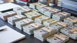 A photo of a close-up shot of a desk covered with multiple stacks of money. There are bills with various denominations, including $1, $5, $20, and $100 notes. The stacks of money are neatly arranged and take up the entire desk. There are also a pen and a notepad on the desk. The background is blurred and contains a few objects.