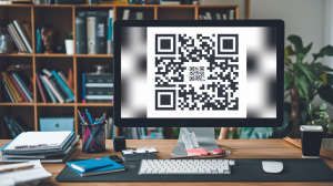 A photo of a wooden desk with a computer monitor on it. The monitor displays a grid of black and white squares. The grid has a QR code in the center. The desk is cluttered with office supplies such as a pen, a notepad, and a ruler. The background is blurred and consists of a bookshelf filled with books and a potted plant.