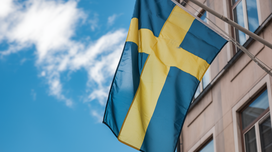 A close-up shot of a blue sky with a few white clouds and a Swedish flag. The flag is hanging from a pole and is fluttering in the breeze. The background contains a building with windows.