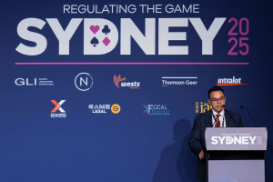 AUSTRAC CEO warns gambling industry to comply with regulations or face tough action. A man in a dark suit, red tie, and glasses speaks at a podium during the 