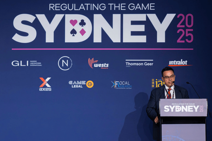 AUSTRAC CEO warns gambling industry to comply with regulations or face tough action. A man in a dark suit, red tie, and glasses speaks at a podium during the "Regulating the Game Sydney 2025" event. The backdrop features the event title in large white letters, with "SYDNEY" stylized to include playing card suits inside the letters. Various sponsor logos, including GLI, Thomson Geer, Intralot, and Game Legal, are displayed on the backdrop. The podium also bears the event's branding. The speaker appears serious and focused.