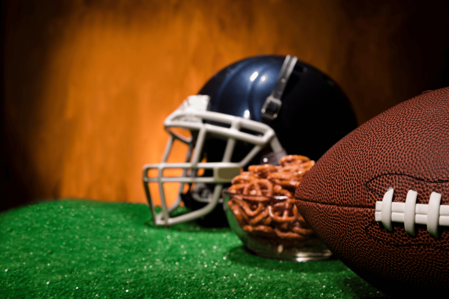 How NFL results could impact gambling giants Flutter and Entain's earnings. A close-up of an American football on artificial turf, with a football helmet and a bowl of pretzels in the background, symbolizing game-day excitement and sports betting.