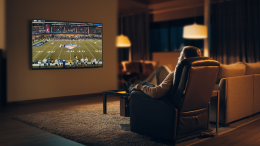 A cinematic shot of a dark room with a person watching a TV mounted on the wall. The TV is displaying an American football game. The person is sitting in a recliner chair. The room has a sofa, a rug, and a table. The background is blurred. The lighting is warm.