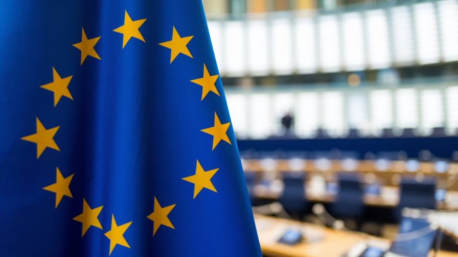 A close-up shot of the European Union flag with a blue background. The flag has a circle of 12 yellow stars on a blue background. The background is blurred, showing a conference room with a table and chairs.