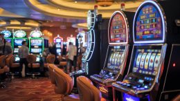A photo of a close-up shot of a casino. There are various slot machines with bright lights and sounds. The background is dark, with a few people standing around. The lighting is warm.