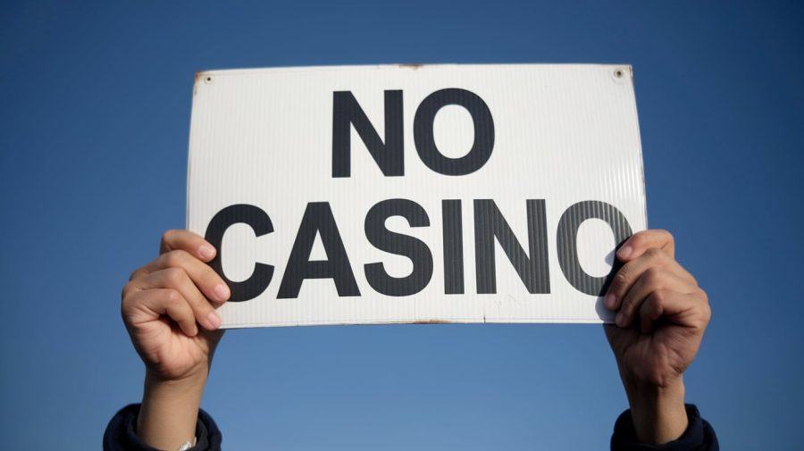 A photo of a person holding up a sign that says 'No casino'. The background is a clear blue sky. The hands holding up the sign are visible.