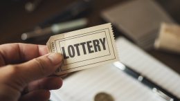A photo of a hand holding a lottery ticket. The ticket has the text "LOTTERY" printed on it. The background is blurred and contains a few other objects, including a pen and a coin. The lighting is soft.