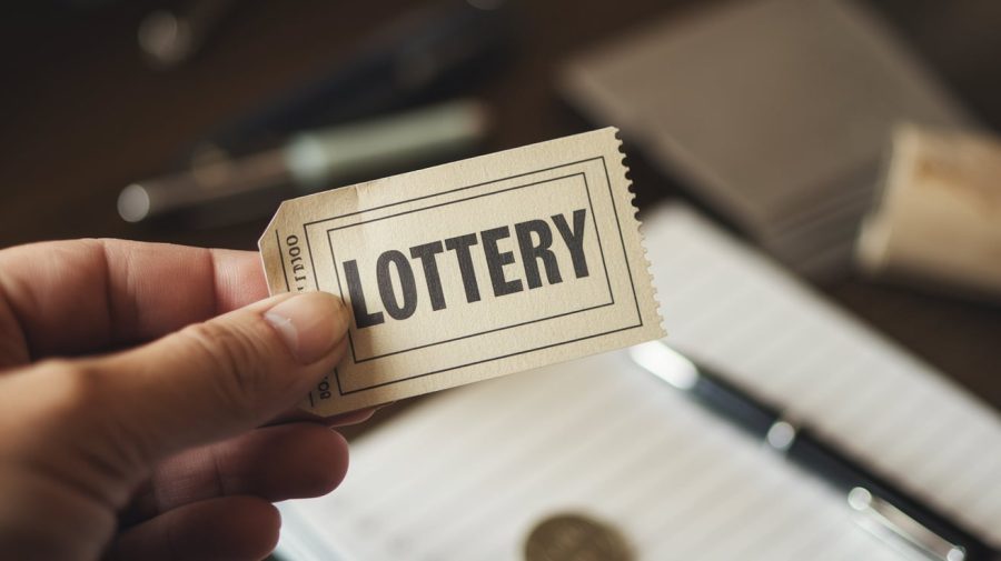 A photo of a hand holding a lottery ticket. The ticket has the text "LOTTERY" printed on it. The background is blurred and contains a few other objects, including a pen and a coin. The lighting is soft.