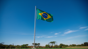 A photo of a clear blue sky with a large Brazil flag waving in the breeze. The flag is placed on a tall mast, and the ground beneath it is covered in green grass. The background contains a few trees.