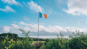 A photo of the Ireland flag flying high against a clear blue sky. The flag is positioned on a tall pole. The background reveals lush greenery. The sky is adorned with fluffy white clouds. The overall scene is serene and picturesque.