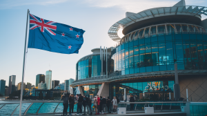 A photo of the New Zealand flag next to a casino. The flag is fluttering in the wind. The casino building has multiple floors and features a modern architecture with a glass facade. There are people standing near the flag. The background contains a city skyline with tall buildings.