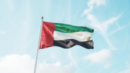 A photograph of the UAE flag billowing in a gentle breeze against a clear, bright blue sky. The flag's vibrant red, white, and green panels ripple gracefully, catching the sunlight. The flag is centered in the frame, with a slight upward angle to emphasize its movement, and the azure sky fills the background with fluffy white clouds. Soft, natural light illuminates the scene, creating a sense of peaceful pride.