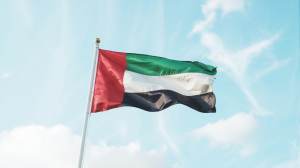 A photograph of the UAE flag billowing in a gentle breeze against a clear, bright blue sky. The flag's vibrant red, white, and green panels ripple gracefully, catching the sunlight. The flag is centered in the frame, with a slight upward angle to emphasize its movement, and the azure sky fills the background with fluffy white clouds. Soft, natural light illuminates the scene, creating a sense of peaceful pride.