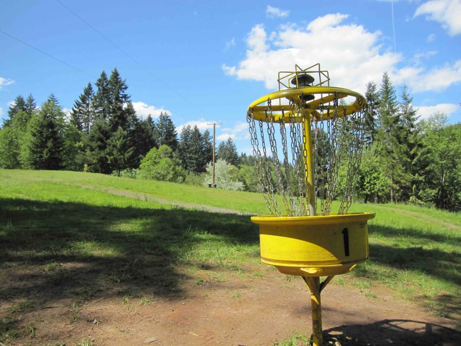 Close-up of a disc golf basket with a solar powered light on it. It's yellow, with a park in the background.