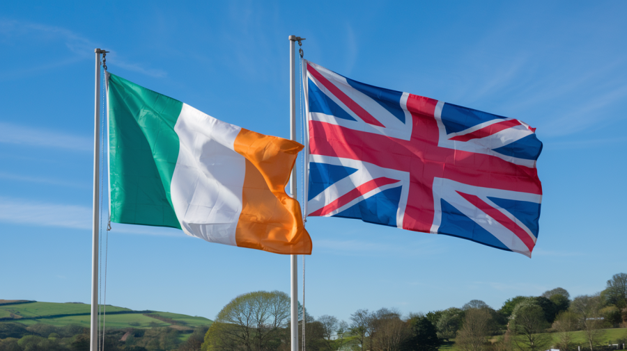 A photo of the Irish flag and the Union Jack flag flying in the wind next to each other. The flags are waving in the breeze against a clear blue sky. The background contains green hills and trees.
