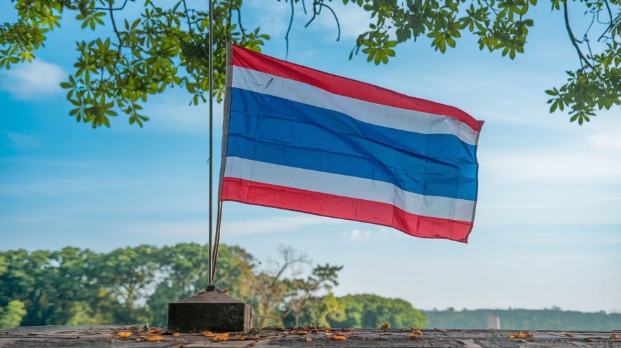 A photo of the Thailand flag waving in the wind. The flag is placed on a pole and is partially unfolded. The background reveals a clear blue sky and lush green trees. The pole is placed on a stone base. There are leaves on the ground around the base. The flag is slightly worn out, with the red and white stripes showing signs of fading.