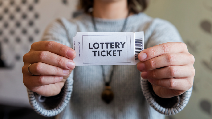A photo of a person holding a ticket that says 'lottery ticket' on it. The person's back is visible, and they are wearing a gray sweater and a pendant necklace. The background is blurred and contains a wall with a pattern.