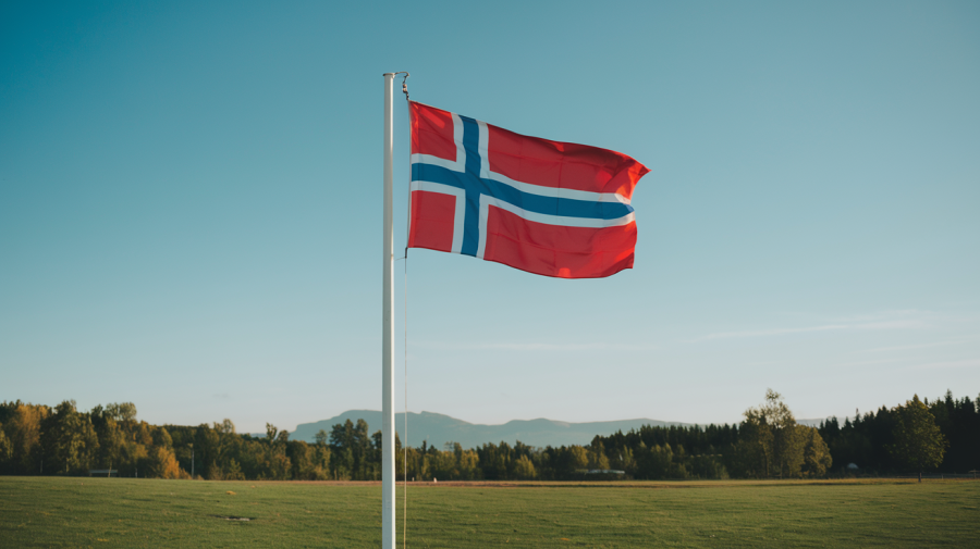 A photo of the Norway flag fluttering in the wind against a clear blue sky. The flag is mounted on a pole standing on a green field. The background is a picturesque landscape with mountains, trees, and a building. The overall image has a serene atmosphere.