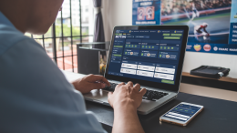 A shot of a person in Thailand betting online. He is sitting at a desk with a laptop. The laptop screen displays a betting website. There is a smartphone next to the laptop. The room has a poster of a sports event on the wall.