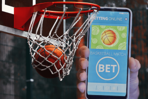 NBA presses CFTC to regulate growing, unchecked sports prediction markets. A basketball going through a hoop with a smartphone in the foreground displaying a sports betting app for a basketball match.