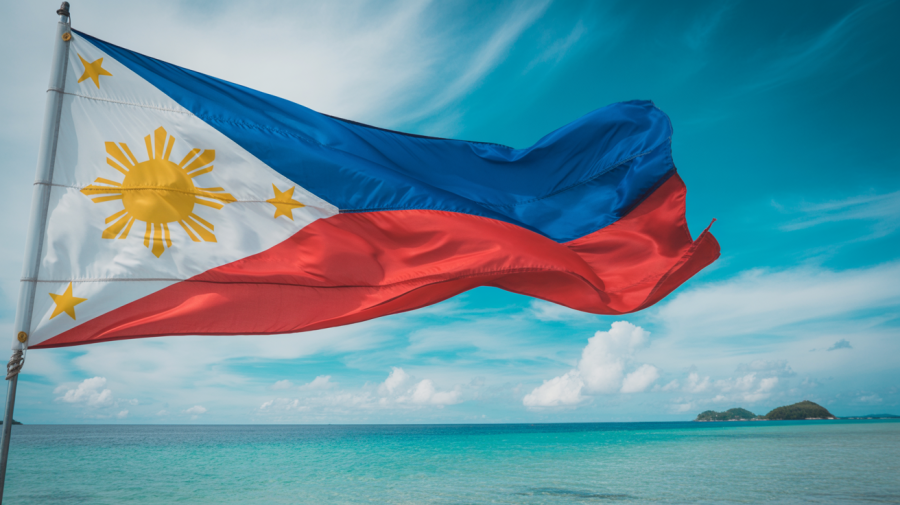 A photograph of the Philippine flag waving proudly against a bright blue sky. The flag’s red and white stripes and yellow triangle ripple dynamically in the wind, showcasing the vibrant colors and intricate design. Below the flag, a tranquil expanse of turquoise ocean stretches towards the horizon with a few small islands visible in the distance. Soft, natural lighting illuminates the scene, creating a sense of serenity and national pride.