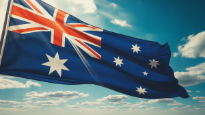 A dynamic, cinematic photograph captures a vibrant Australian flag rippling powerfully in a strong wind. The flag's deep blue background and the bright red stars and Union Jack section are sharply defined against the azure sky, exhibiting a sense of patriotic movement. Sunbeams cut through the clouds, casting dramatic light and shadows across the flag’s fabric, highlighting its texture and creating a visually arresting effect. The background consists of a clear, expansive blue sky with scattered fluffy white clouds, emphasizing the flag’s prominence and conveying a feeling of open space and boundless freedom.
