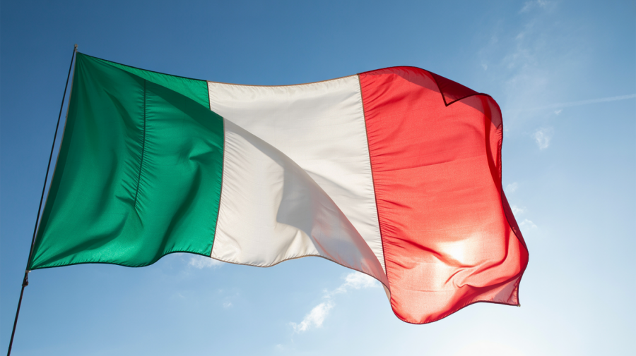 A photograph of an Italian flag billowing in a gentle breeze against a clear blue sky. The flag's vibrant red, white, and green stripes ripple dynamically, catching the sunlight as they wave. The sky is a brilliant azure, with a few wispy clouds scattered across the upper portion of the frame. Soft, diffused lighting emphasizes the flag's colors and creates a sense of movement.
