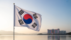 A photograph of the South Korean flag waving gently in a light breeze against a clear blue sky. The flag's vibrant colors—white, blue, and red—are crisp and distinct, with the Taegeuk symbol perfectly centered. In the distant background, blurred and partially obscured by a light haze, sits a glittering casino complex with its neon lights reflecting off a calm body of water. Soft, diffused sunlight illuminates the flag and the scene, creating a sense of serene grandeur.