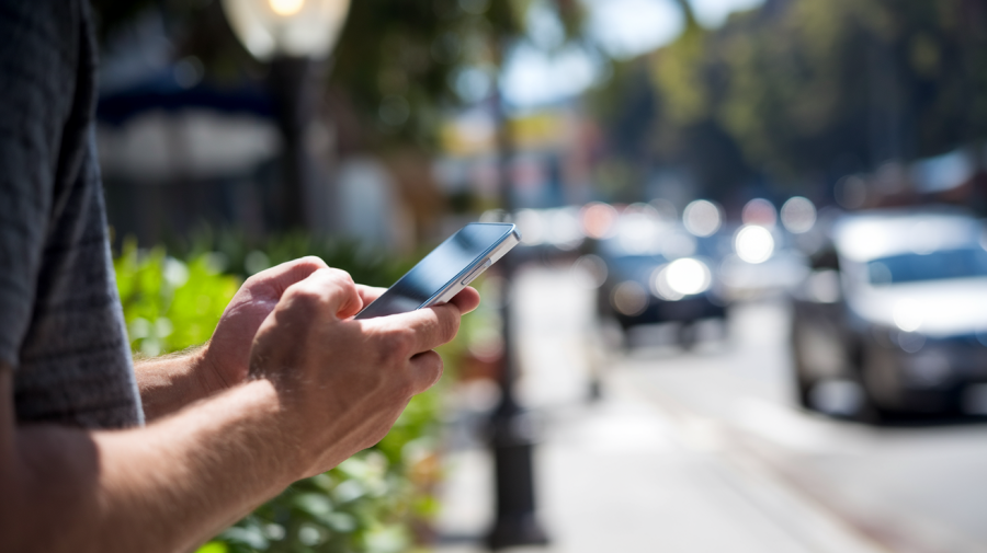 A photo of the back of a man holding a phone in his hands. The man is standing on a sidewalk with green plants and a lamp post behind him. The background is blurred, showing a city street with cars. The lighting is bright.