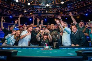 Poker table with people crowded around in the background. All celebrating with arms in the air.