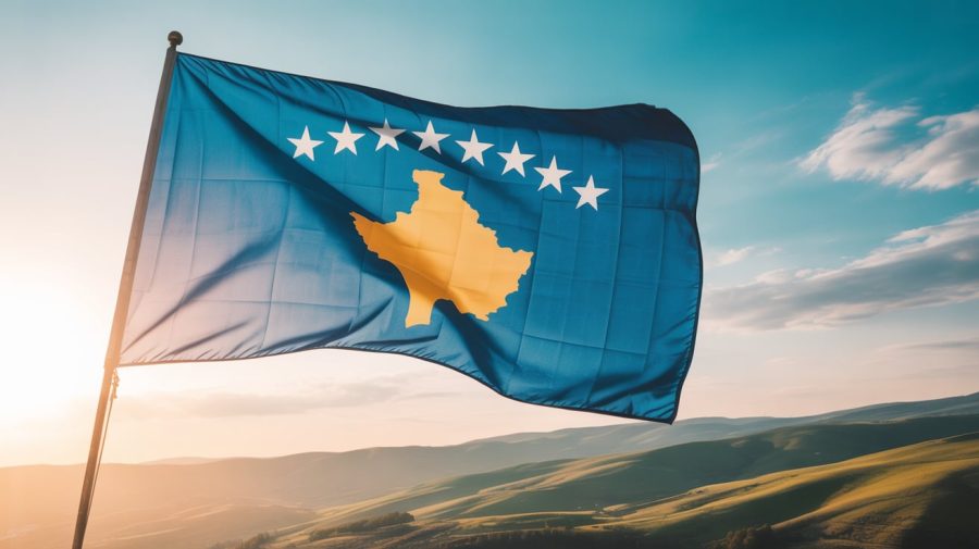 A dramatic, cinematic photograph of the Kosovo flag waving proudly in a gentle breeze. The flag's vibrant red and yellow colors are sharply defined against a clear, azure sky, with the fabric rippling and flowing dynamically. Below the flag, rolling green hills stretch into the distance, suggesting the landscape of Kosovo, bathed in the warm light of a late afternoon sun. The composition emphasizes the flag's movement and symbolism, evoking a sense of national pride and resilience.