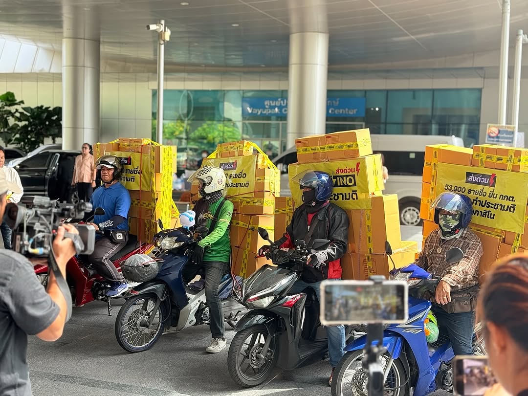 Motorcycle delivery riders carrying stacks of yellow boxes labeled with protest signs, including “ส่งด่วน NO CASINO ต้องประชามติ” and “53,900 รายชื่อ ยื่น กกต. ทำประชามติ หยุดกาสิโน,” outside a Thai government building as they hand in petition over referendum.