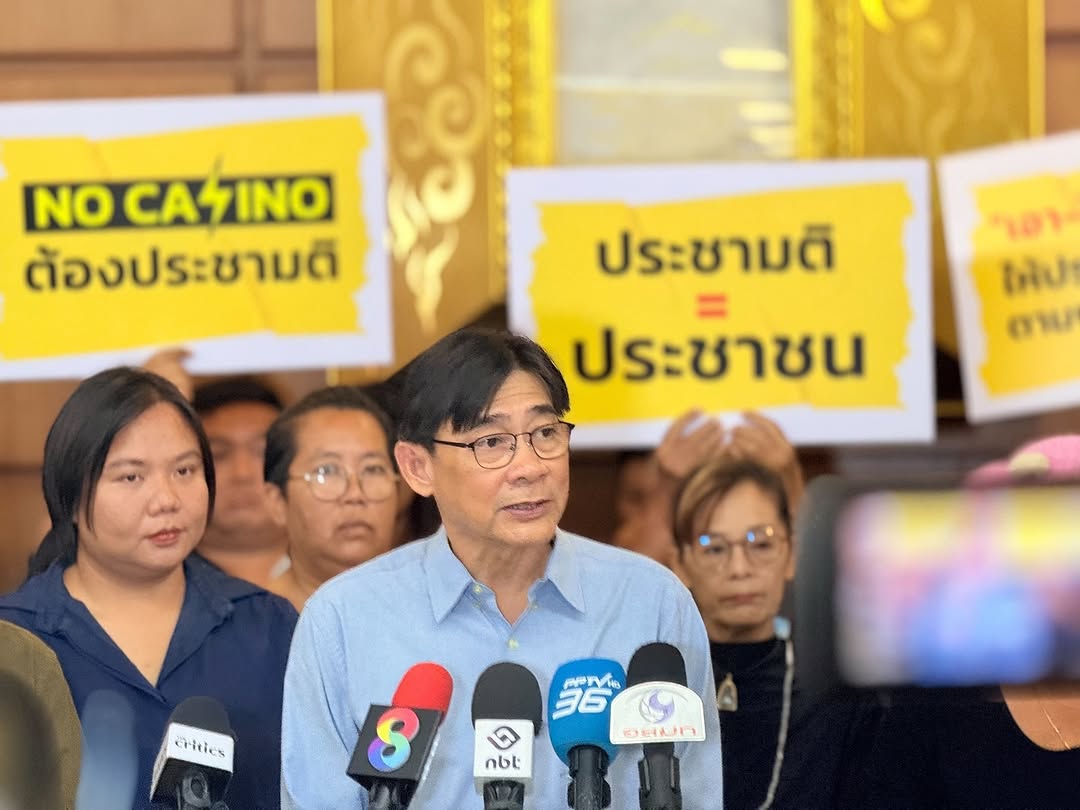 A man speaks at a press conference surrounded by supporters holding yellow protest signs. Prominent signs behind him read “NO CASINO ต้องประชามติ” and “ประชามติ = ประชาชน” in Thai.