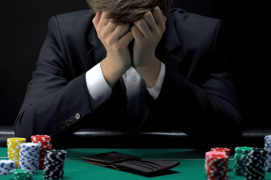 Gambling tops digital addiction risks in 2025, especially for young men - study. A man in a suit sits at a poker table with his head in his hands, appearing distressed. Poker chips and an empty wallet lie in front of him, suggesting financial loss from gambling.