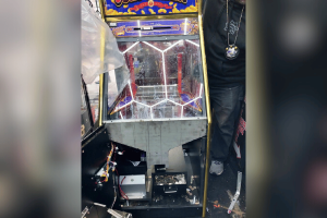 Illegal gambling machines seized in San Francisco drug bust. A dismantled illegal gambling machine with its interior components exposed, including wires, coins, and mechanical parts. A person stands next to it, partially visible, wearing dark clothing and a medallion. The top of the machine displays colorful graphics and text, indicating it was used for coin-based gambling.