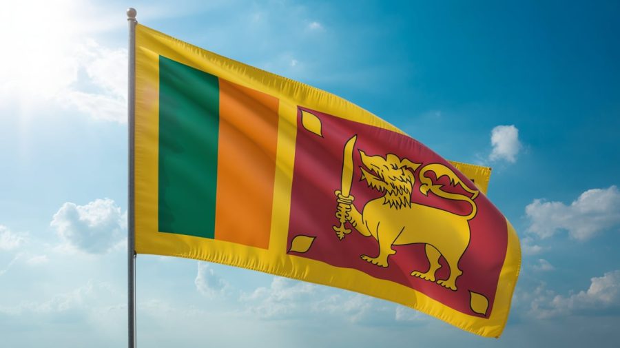 A photograph of the Sri Lanka flag, vividly displaying the vibrant saffron, green, and crimson colors, waving powerfully in a strong wind. The flag is positioned against a clear blue sky, with a few fluffy white clouds scattered in the distance. Sunlight illuminates the flag, creating a dramatic rippling effect and emphasizing the distinct shades of the colors. The scene conveys a sense of national pride and freedom with the flag billowing proudly.