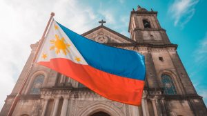 A picture of the vibrant Philippine flag gently waving in the foreground against the backdrop of the Great Historic Catholic Church. The flag is bright, where the sun is shown prominently in the left upper corner, fluffing a little in a gentle breeze. The church, made of roaming stone, contains complex sculptures and stained glass windows, with a tall bell tower that reaches a clear blue sky. The soft natural light lights the scene, highlights the strength of science and the facade of the church.