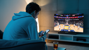 A photograph of the back of a young man wearing a grey hoodie, his head slightly bowed as he intently scrolls through his phone. He is seated on a plush, dark grey sofa in a modern living room, facing a large television displaying a fast-paced basketball game. The room is dimly lit by a soft glow emanating from the TV screen and a minimalist floor lamp, casting subtle shadows across the textured wall behind him. A half-empty glass of iced tea rests on the coffee table in front of the sofa.