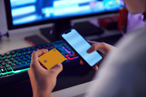 Gambling addict jailed after stealing $2.3M from recruitment firm employer. Person holding a yellow credit card and a smartphone in front of a gaming keyboard and computer screen, suggesting online activity or digital transactions.