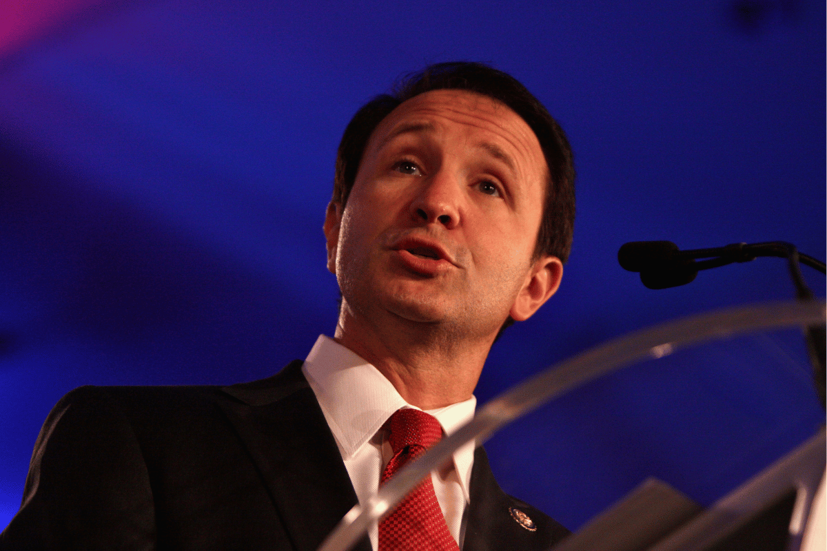 Governor Jeff Landry speaking at a podium, wearing a dark suit and red tie, with a microphone in front of him against a blue-lit background.