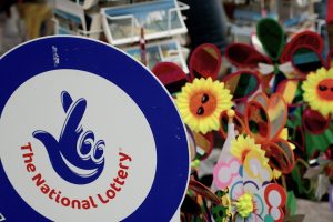 A sign featuring "The National Lottery" logo with a blue background and a hand symbol with crossed fingers. In the background, there are colorful pinwheels and sunflowers with smiling faces, likely part of a festive street stall or market. UK's National Lottery to temporarily shut down for major tech updates