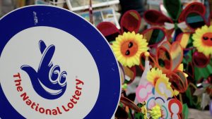 Mark "National lottery" A blue background logo and a code with cross fingers. In the background, there are colored cupboards and sunflower flowers with smiling faces, which are likely to be part of a stall or festive market in the street. The national lottery will be closed in the UK temporarily due to the main technical updates. The national lottery in the United Kingdom dates back to the slogan of the nineties 